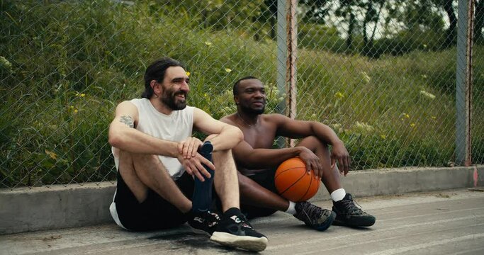 Two men are sitting on the floor of the basketball court and chatting about the ongoing match, they are happy that they can attend the game