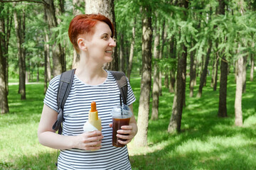 middle-aged woman holds a hot dog in her hands and cold black coffee in a plastic cup. Snack during a walk in the city park.