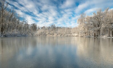 Landscape of a frozen lake surrounded by trees and snow under a blue cloudy sky