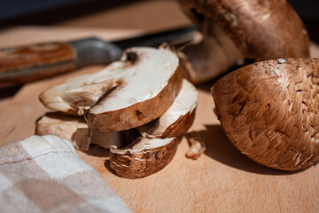 mushrooms (large champignons with a brown cap) lie on a wooden board, one mushroom is cut and the other is whole, a knife and a kitchen towel are next to it