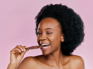 Black woman, eating chocolate and diet with candy and beauty, health and dessert snack on pink background. Skin, glow and female model wink with cacao sweets, calories and cosmetics in studio