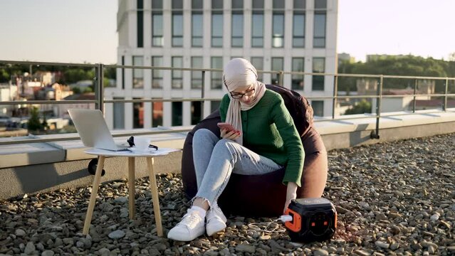 Full length of muslim woman plugging mobile charger into wall outlet of portable power station on panoramic terrace. Relaxed lady in headscarf activating digital device for making business calls.