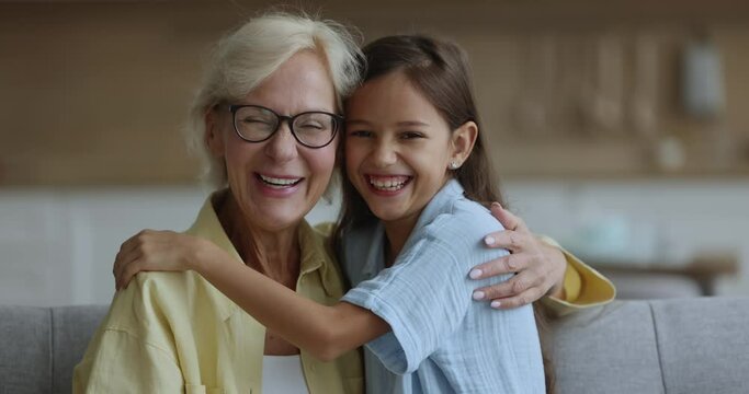 Close up portrait happy granddaughter hugs older carefree grandma sit on couch at living room, look camera, pose, spend time together, two age generations enjoy weekend at home feel unconditional love