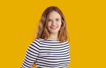 Studio portrait of pretty woman. Headshot of happy beautiful young blonde long haired girl in casual striped top standing isolated on orange yellow background, looking at camera and smiling