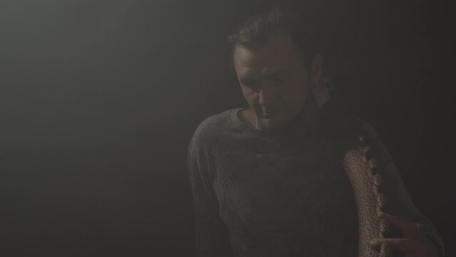 A hipster man sits on the floor and holds an Australian aboriginal wind musical instrument Didgeridoo on a dark background. The camera is moving