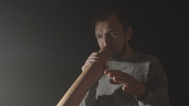 A hipster man plays the Australian aboriginal wind musical instrument Didgeridoo on a dark background. Music footage. Studio photography