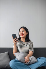 Asian woman using smartphone while laying on the sofa at home. Cheerful lady enjoys online chatting, spends time in social networks, looks at the phone and smiles