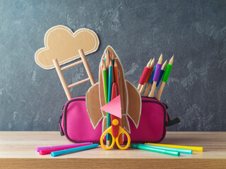 Back to school concept with rocket, pencils and pink pencil case on wooden table over blackboard...