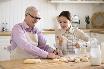 Positive teenage girl in apron helping grandpa to cook in home kitchen, sharing domestic chores, preparing homemade bakery meal, shaping, stretching, rolling dough at floury table