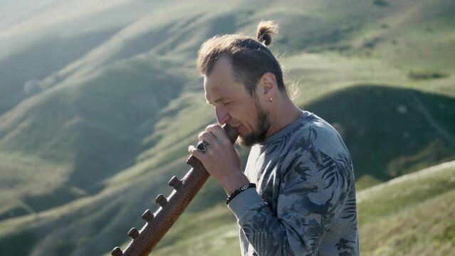 The musician plays the Didgeridoo against the backdrop of hills with feather grass. Bright sunny day. The camera is moving