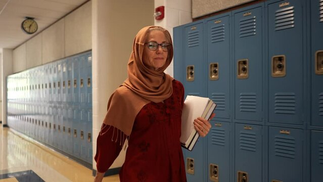 Slow motion diagonal front view of happy smiling muslim teacher wearing headscarf walking down a hallway in an empty school holding books with US flag in background.