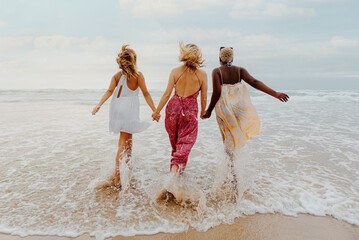 view from behind of multiracial group of young women on the shore of the beach with their hands...