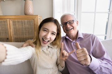 Self portrait of happy excited teenager granddaughter kid and cheerful older grandpa showing hand symbols, gestures at camera, smiling, laughing, having fun, taking selfie together at home