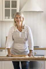 Happy pretty blonde mature baker woman in apron rolling raw dough on wooden rustic kitchen table, baking fresh homemade buns, looking at camera, smiling. Vertical shot portrait