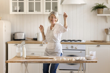 Cheerful active pretty mature woman dancing at floury kitchen table with bakery food products,...