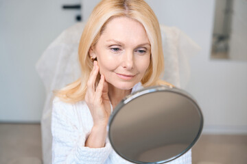 Happy mature lady looking in mirror at cosmetologist office