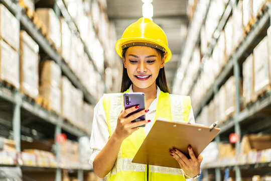 Young asian woman worker uses the phone for business contacts and holding clipboard to checking inventory in the warehouse - Powered by Adobe