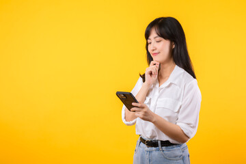 Portrait beautiful asian young woman wearing white shirt and denim plants thoughtful body gesture with smartphone isolate on yellow studio background. Thinking positive in online news concept.