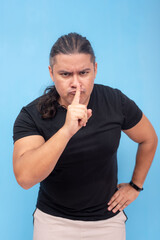 An irate biracial male leaning forward and doing a shush gesture with one finger. Isolated on a blue background. - obrazy, fototapety, plakaty