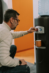 A middle-aged man pouring hot water from the water dispenser in the cup. He is preparing tea