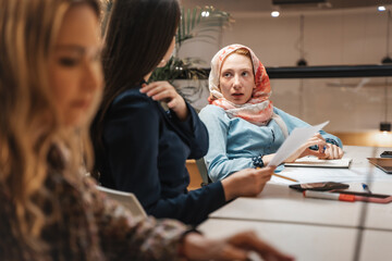 Female employee wearing hijab listening to her brunette coworker while working together at the office