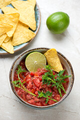 Bowl of tomato salsa served with nachos chips, vertical shot on a light-beige stone background, selective focus