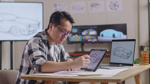 Side View Of Asian Male Looking At The Photo On Paper Then Drawing New Car Design Concept On A Tablet In The Studio With Tv And Computers Display 3D Electric Car Model 
