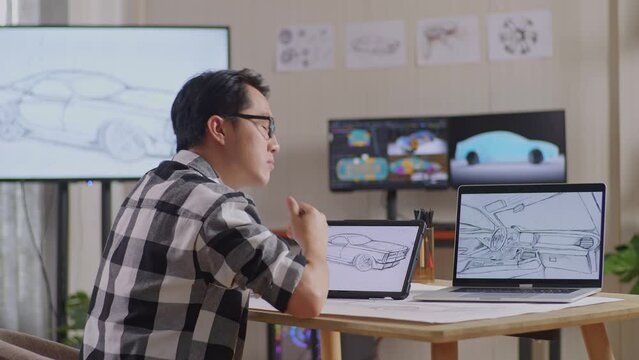Side View Of Asian Male Thinking About New Car Concept Then Raising Index Finger While Drawing On Tablet In The Studio With Tv And Computers Display 3D Electric Car Model 
