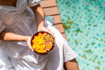 Obraz na płótnie Canvas Young woman traveller relaxing and eating refresh bowl with fruits at tropical resort pool on summer vacation
