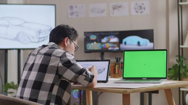 Side View Of Asian Male Having A Headache While Drawing Car Concept On Tablet Beside Green Screen Laptop In The Studio With Tv And Computers Display 3D Electric Car Model 
