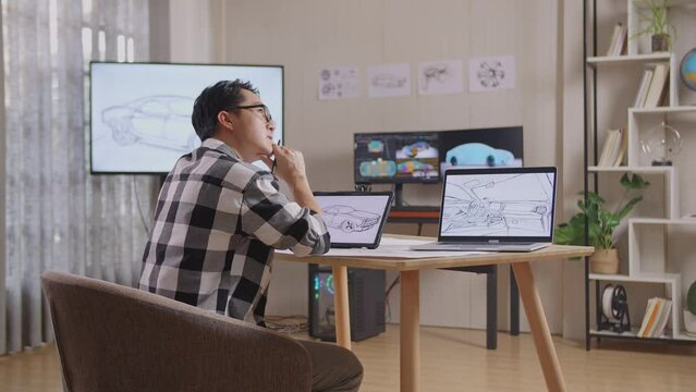 Side View Of Asian Male Thinking About New Car Concept While Working On A Car Design Sketch On Tablet In The Studio With Tv And Computers Display 3D Electric Car Model 
