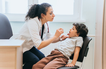 Serious brunette Spanish female doctor checks throat of little  boy at medical office. Pandemic concept. Toddler at medical exam at hospital. Healthcare, healthy  childhood.