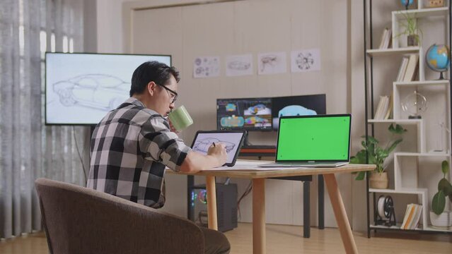 Side View Of Asian Male Drinking Coffee While Drawing Car Concept On Tablet Beside Green Screen Laptop In The Studio With Tv And Computers Display 3D Electric Car Model 

