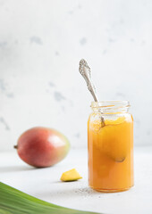 Glass jar with spoon of fresh organic mango jam marmalade puree on light background.