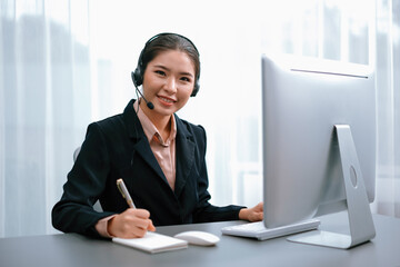 Asian call center with headset and microphone working on her workspace looking at camera. Female...