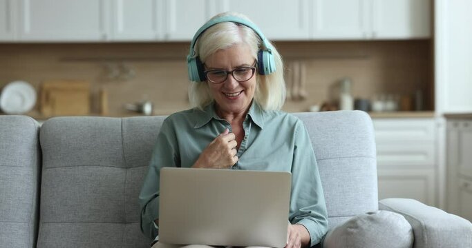 Cheerful positive elder blonde lady in trendy headphones waving friendly greeting hand hello at laptop, speaking on online video conference call, using hand gestures, body language