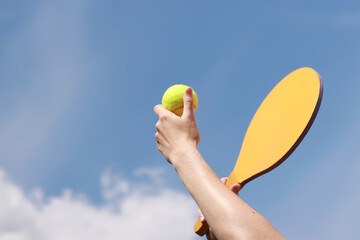 Play paddleball with a wooden racket against a blue sky. Beach tennis. A wooden paddle for playing tennis and a ball in the hands of a girl. Outdoor games. Close-up
