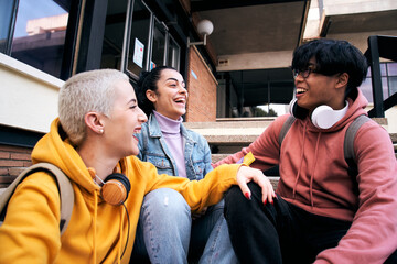 Group of three students laugh a lot sitting outdoors. Teenage gen z multiracial classmates are...