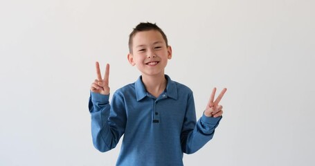 Asian schoolboy displays the iconic Victory gesture on white backdrop. With a beaming smile and outstretched fingers forming the victory sign, this young boy radiates positivity and enthusiasm.