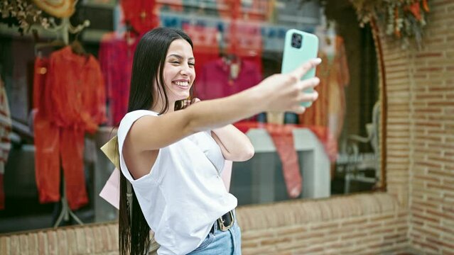 Young beautiful hispanic woman make selfie by smartphone holding shopping bags at clothing store