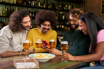 Group of young people watching at mobile in a bar. This four multi-ethnic friends are smiling and having fun while they look at a smartphone. Millennial guys using a cell indoors. Technology concept