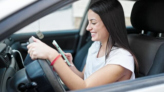 Young beautiful hispanic woman using smartphone sitting on car at street