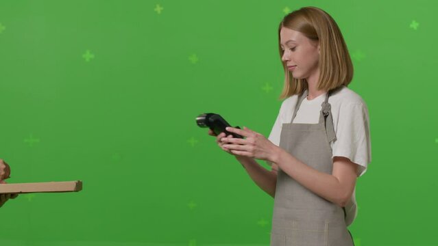 Girl waiter passing a box of pizza on green screen chroma key background. Woman hold wireless bank payment POS terminal to process acquire credit card payment. Copy space mock up advertising