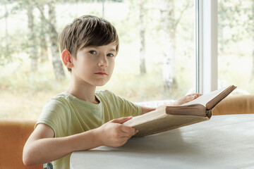 A child sits at the table and reads an old book. A boy with a book in his hand. A child's lesson with a book