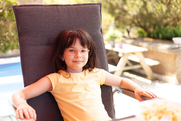 Naklejka premium Cheerful little laughing girl in glasses sits in a chair in a cafe on a warm summer day