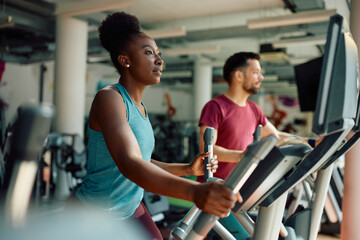 Young black sportswoman using elliptical trainer while exercising in gym. - obrazy, fototapety, plakaty