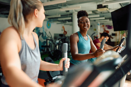 Happy black athlete and her friend exercising on cross trainer during sports training in gym.