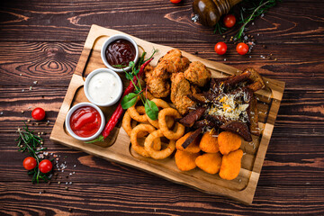Snacks breaded squid rings, chicken nuggets, rye bread croutons with cheese, chicken legs and sauces on a wooden board.