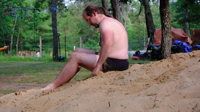 Obese young male wearing only black briefs playing while sitting on pile of sand or sandbox.