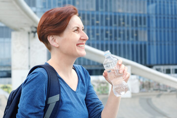 Middle-aged woman with a short haircut drinks water from a plastic bottle against the backdrop of a modern urban business district. The need to drink fluids while in the heat.